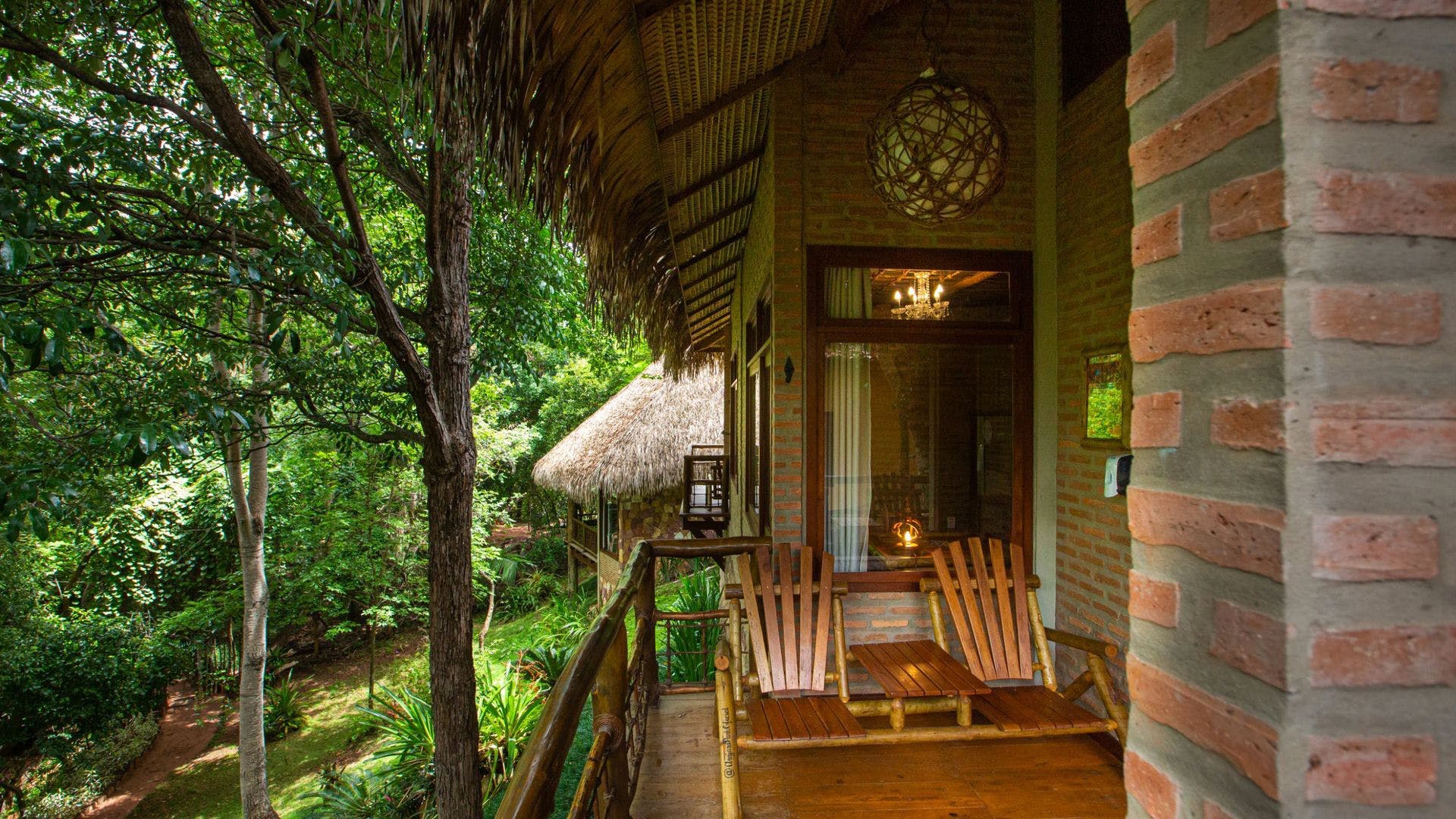 Foto da pousada, onde é possível ver uma varanda que possuí duas cadeiras de bambu, e no lado de fora algumas arvores